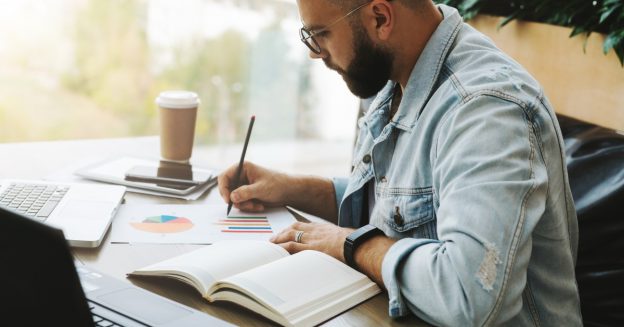 Male, graduate-aged student sitting at table with laptop, coffee and study papers