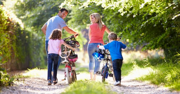 A mother, father and two similarly-aged children (boy & girl) walking their mountain bikes in a gravel road
