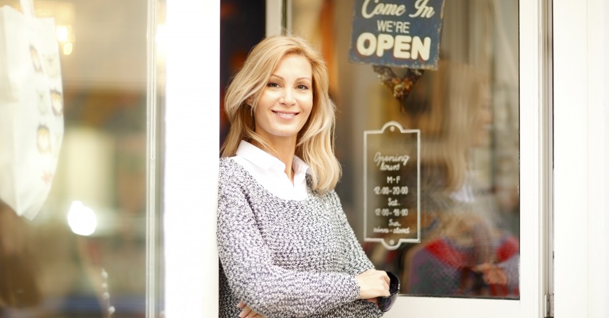 Smiling woman standing in doorway of a business location with business hours posted on signs in the window