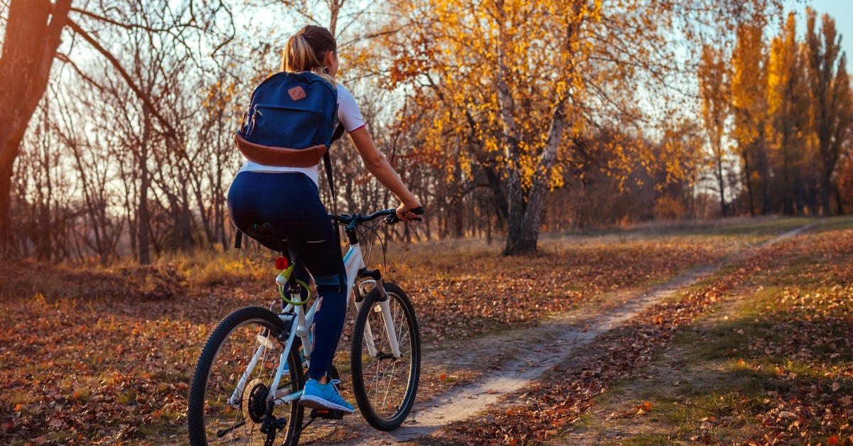 A woman riding a bicycle along a dirt trail beside trees with autumn-yellow leaves