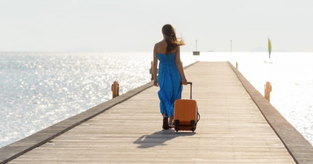 Woman in blue dress walking on a pier traiuling a roller suitcase