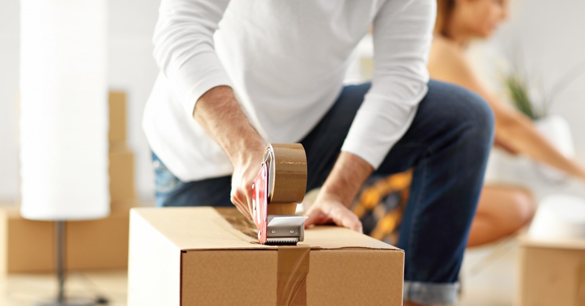 Close-up of man using packing tape to seal a cardboard box with woman in background.