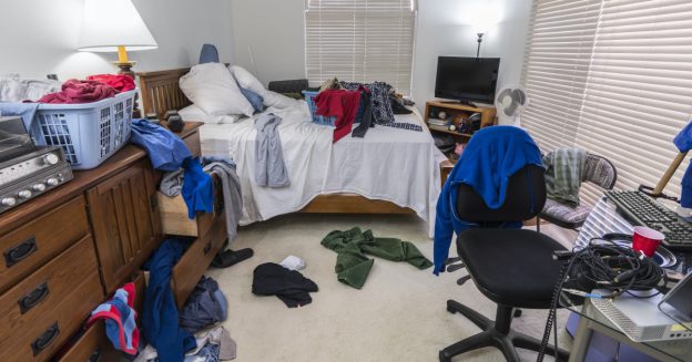 Scene of messy bedroom with laundry strewn about and desk with keyboard.