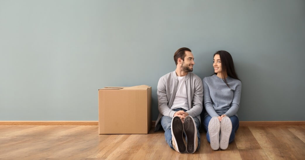 Man and woman seated on floor of empty apartment with moving box next to them.