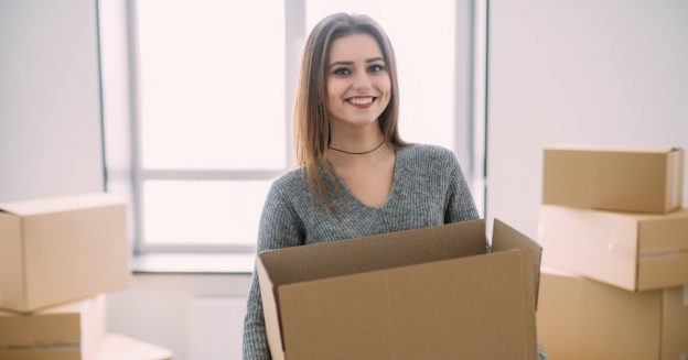 Young woman smiling at camera holding a moving box in a room