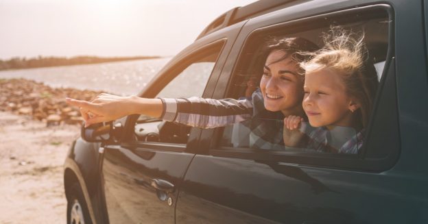 Woman and young girl looking out of the back passenger window of a car and smiling, with beach in background