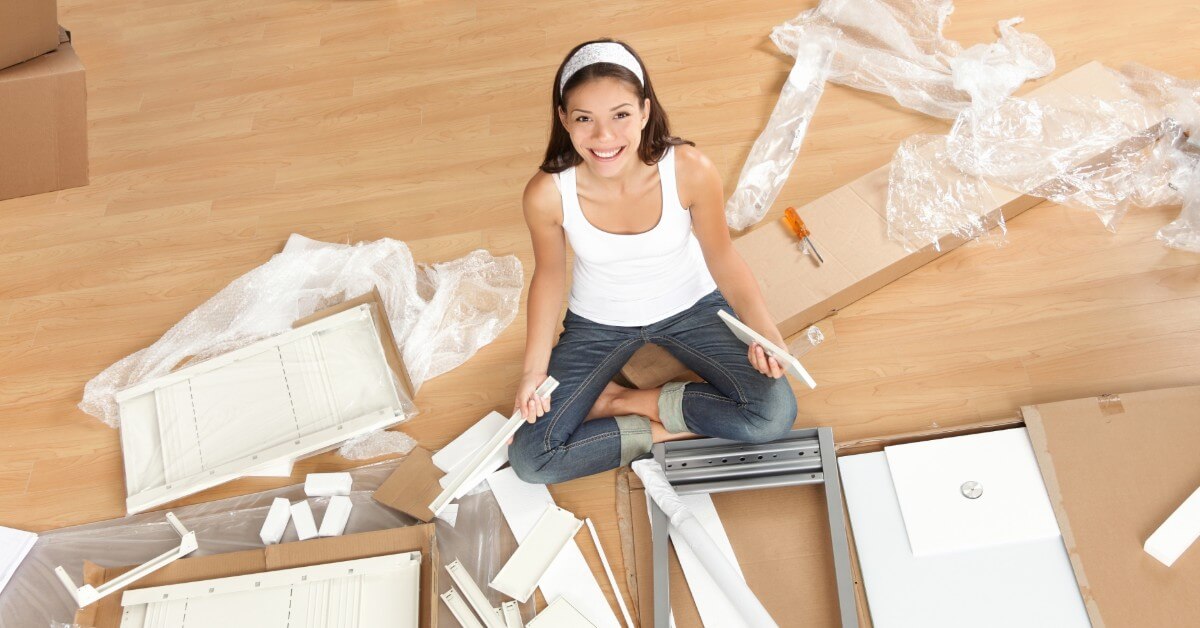 Smiling woman seated on floor looking up while assembling a newly unpacked furniture item.